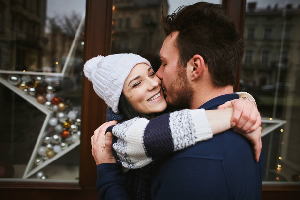 happy enamored couple is standing outdoors near the decorated shop windows, hugging and kissing each other - 写真・画像