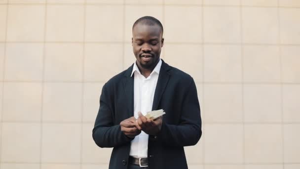 Happy african american businessman holding a big amount of money in his hands. He stands in the street near the office center or bank and shows money to the camera - Video, Çekim