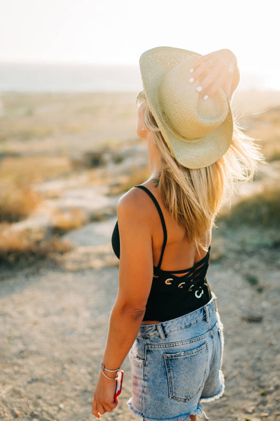 Back view. Fashion portrait of pretty young woman wearing hat and jeans on the beach at sunset - 写真・画像