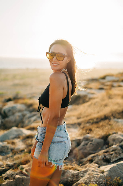 Fashion portrait of pretty young woman with sunglasses and jeans on the beach at sunset, retro style. - Foto, Imagen
