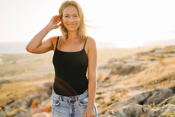 Fashion portrait of pretty relaxed woman with jeans on the beach at sunset, retro and melancholic style. - Foto, imagen