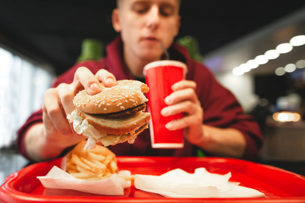Młody człowiek dania fast food w restauracji typu fast food. Człowiek posiada burger w ręku, picie cola z czerwonego szkła. Frytki i taca fast food. Koncepcja Fast Food. Nastolatek zjada fast food - Zdjęcie, obraz