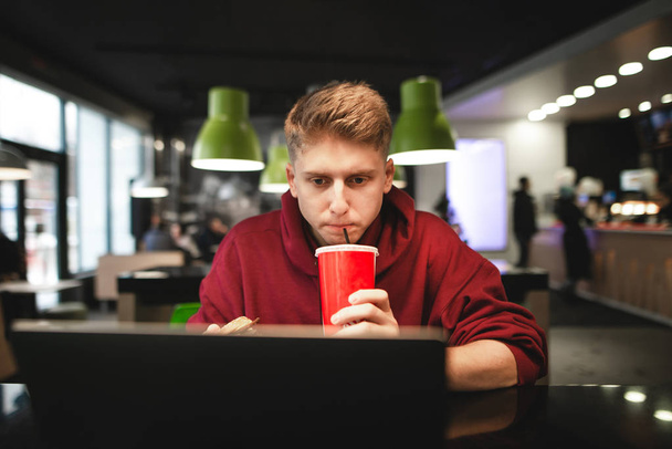 Portrait of a young man using a laptop in a cafe and drinking a red beverage drink. Freelancer working on a laptop in a cafe on the screen looks focused and drinking cola. Work in a cafe - Photo, Image