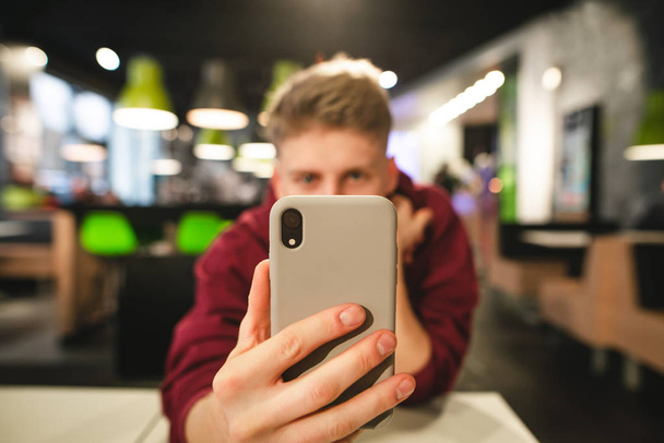 Guy holds the smartphone in his hand at his background. Positive young man takes a photo on a smartphone in a fast-food restaurant, a smartphone in focus close-up in the foreground. Copyspace - Photo, Image