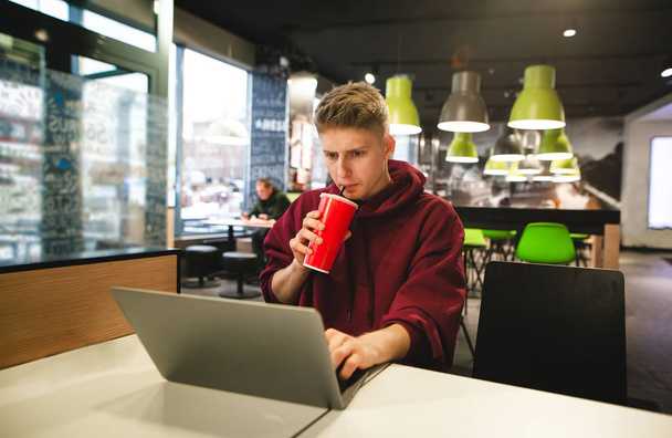 Занятой парень в повседневной одежде работает в кафе на ноутбуке, фокусируется на просмотре экрана и выпивает напиток из бокала. Фрилансер работает в кафе быстрого питания
. - Фото, изображение