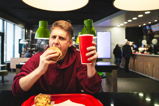 Młody człowiek siedzi w restauracji fast food z tacą z jedzeniem, przytula wielkiego apetycznego hamburgera i patrzy w kamerę. Portret ucznia jedzącego posiłki w restauracji fast food. Nastolatek je hamburgera.. - Zdjęcie, obraz