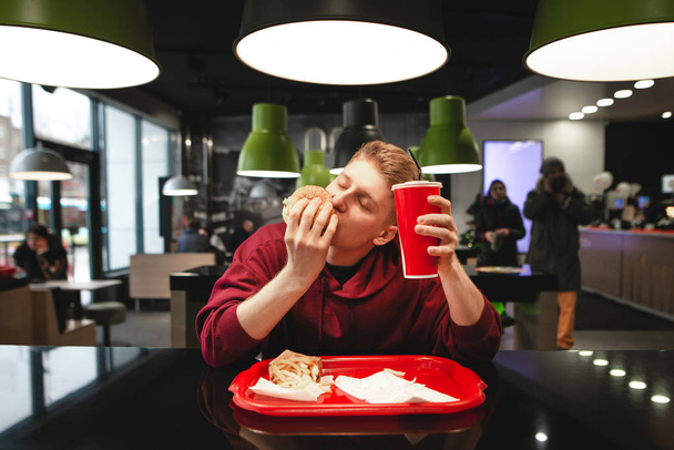 Éhes fiatalember ül az asztalnál egy gyorsétteremben, és megharap egy hamburgert csukott szemmel, egy pohár italt tart a kezében, és örömet szerez. A gyorskaja egy ebéd.. - Fotó, kép