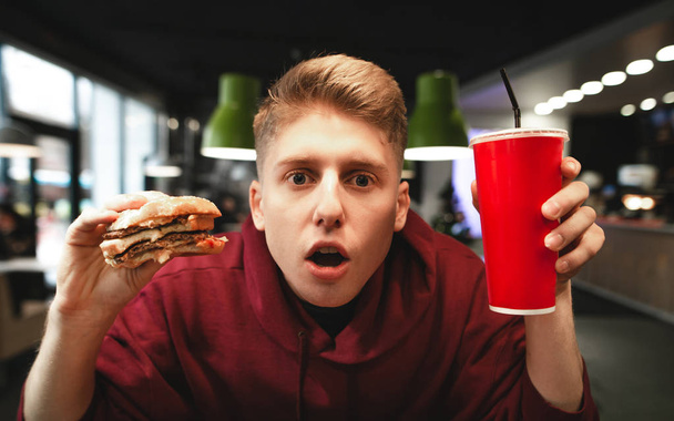 Egy meglepett fiatalember arcképe egy étvágygerjesztő hamburgert és egy pohár italt tart a kezében, belenéz a kamerába. Lenyűgöző ember gyorsétterem eszik a kezében az étterem háttér. - Fotó, kép