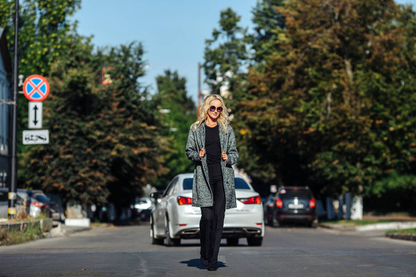 Beautiful blonde girl in a gray coat is walking on the roadway on the background of cars - Photo, Image