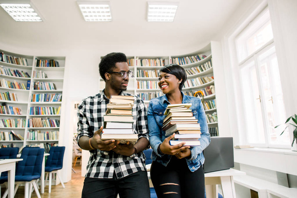 Young smiling african boy and girl holding books standing together on the background of shelves with books in library. Students are studying in library. Academic Library Student Learning Concept - Valokuva, kuva