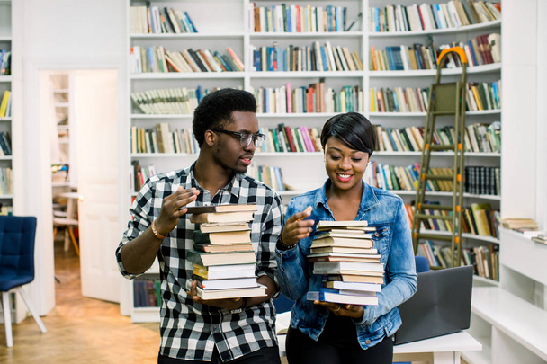 Hipster jonge Afro-Amerikaanse studenten jongen en meisje die in de bibliotheek aan de universiteit studeren. Gelukkig lachend paar bedrijf boeken en plezier in moderne bibliotheek. - Foto, afbeelding