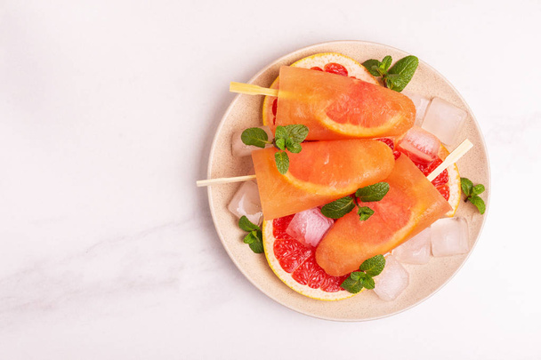 Homemade grapefruit popsicle with ripe grapefruit slices and fresh mint on light marble background. Healthy desserts. Lactose free ice cream. - 写真・画像
