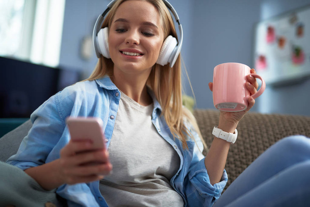 Cheerful pretty girl with hot drink listening music and using cellphone - Foto, Imagem