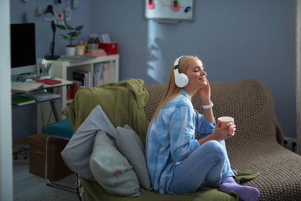 Charming young woman with hot drink listening music at home - Photo, Image