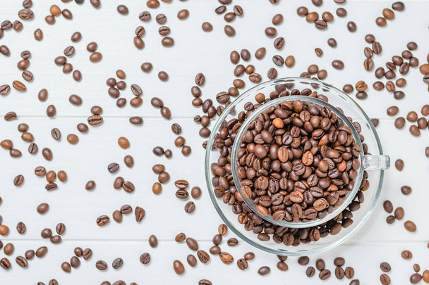 Glass bowl filled with coffee beans on a background of scattered grains on a white table. The view from the top. Flat lay. Grains for the preparation of the popular drink. - Фото, зображення