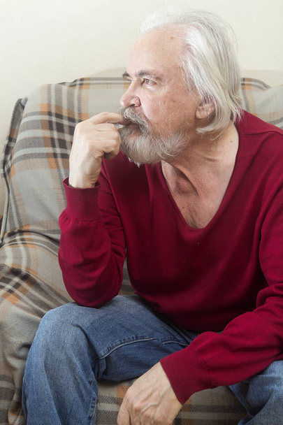 Ένας όμορφος άρρωστος παλιά άνδρας με μούσι και μουστάκι με μακριά λευκά μαλλιά σε γηροκομείο κάθεται σε μια πολυθρόνα ολομόναχος και αντανακλά τα τελευταία χρόνια - Φωτογραφία, εικόνα