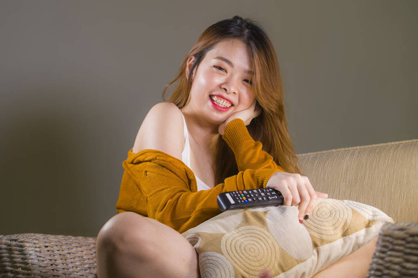 young happy and attractive Asian Korean woman watching television at home sofa couch holding remote smiling relaxed enjoying comedy movie or TV show in domestic entertainment concept - Фото, зображення