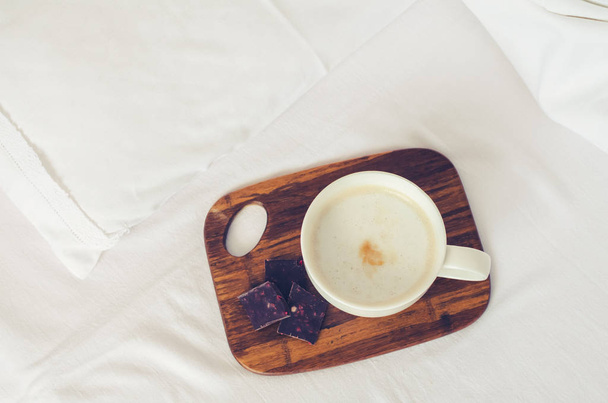 Cup of coffee cappuccino with dark chocolate on wooden board on a bed on cozy lazy sunday. White bedding sheet, blanket and pillows. Good morning concept. Enjoy slow life. Top view. - Photo, Image
