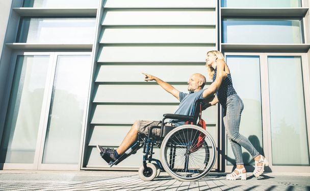 Szczęśliwa para z niepełnosprawny mężczyzna kręcącego się w miejskich miasta tło - koncepcja relacji w kwestii niepełnosprawności z facetem na wózek inwalidzki i młody asystent kobieta - jasne desaturated filtr - Zdjęcie, obraz