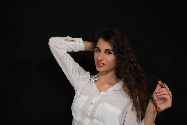 Young beautiful girl with long wavy brown hair and arms in her hair. He wears a white men's shirt, a smiling and sensual attitude. Half-length portrait. Black background. Copy space - Φωτογραφία, εικόνα