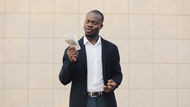 Portrait of african american businessman in suit counting money standing near office centre. He celebrating his successful win with a lot of dollars and looking into the camera - Séquence, vidéo