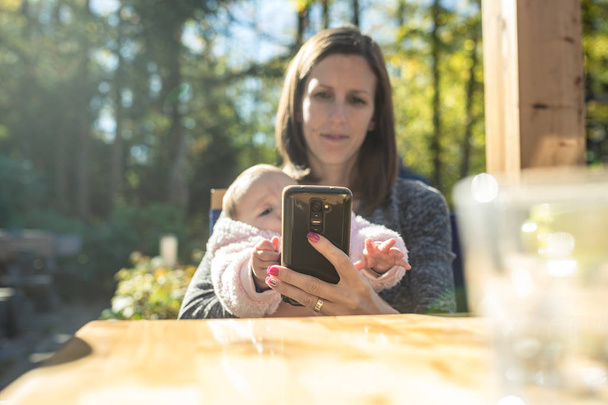 Νεαρή μητέρα χρησιμοποιώντας το κινητό της τηλέφωνο, όπως κάθεται έξω από ένα καφέ κρατώντας την κόρη του μωρού φθάνει για το τηλέφωνο. - Φωτογραφία, εικόνα