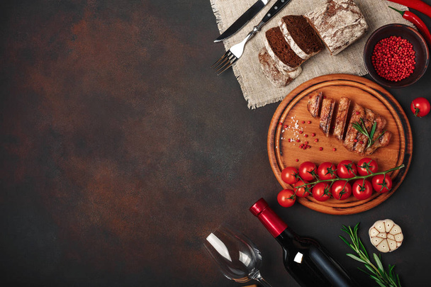 Sliced grilled pork steaks with bottle of wine, wine glass, corkscrew, knife, fork, black bread, cherry tomatoes, garlic, onion and rosemary on rusty background. Top view. - Photo, Image