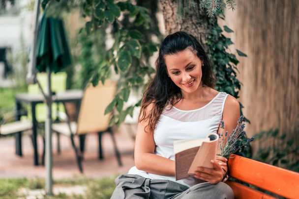 Beautiful young woman reading a book on the bench outdoors. - 写真・画像