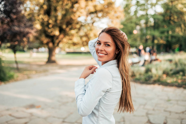 Venkovní portrét usměvavé mladé ženy. Dívka stojící v městském parku, při pohledu na fotoaparát. - Fotografie, Obrázek