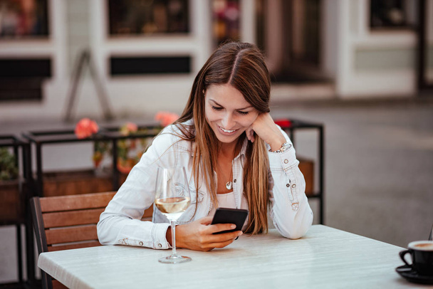Szczęśliwa młoda kobieta siedzi w restauracji na świeżym powietrzu, patrząc na smartfonie. Kieliszek białego wina na stole. - Zdjęcie, obraz