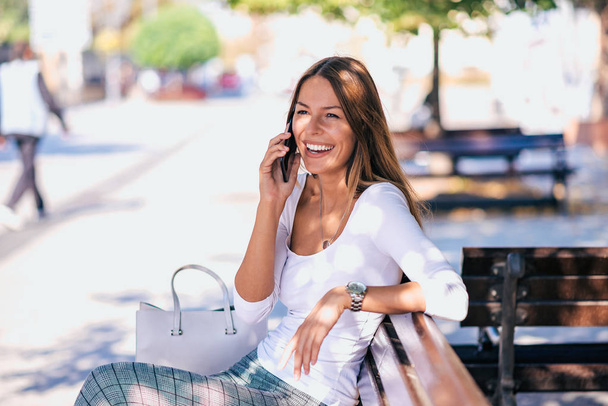 Happy young woman sitting on a bench and talking on the phone. - Photo, image