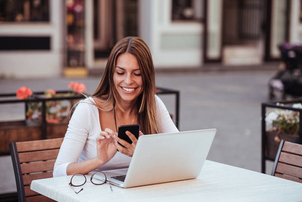 Portrait of a smiling young woman using mobile phone while sitting with laptop at a cafe outdoors. - Photo, image