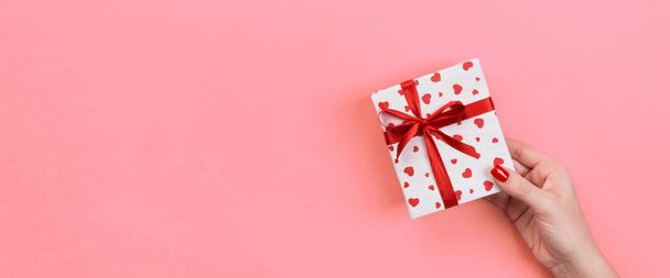 女性の手は、紙に赤いリボンでバレンタインまたは他の休日の手作りプレゼントを与えます。コピー スペースを設計するためにサンゴ色のテーブルの上のギフト、トップ ビュー バナーのボックス、赤い心装飾を提示します。. - 写真・画像