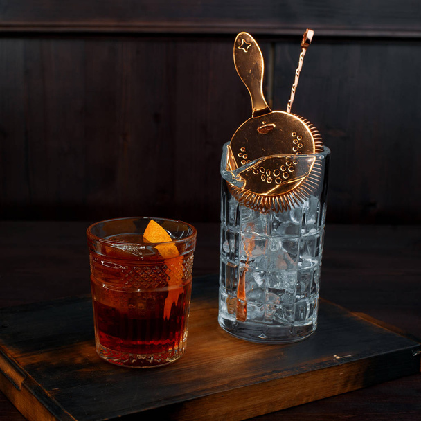 Whisky con una rebanada de naranja en un vaso de cristal está sobre la mesa en el bar junto al vaso con hielo y herramienta profesional para hacer un cóctel. Bebida fuerte especiada
. - Foto, imagen