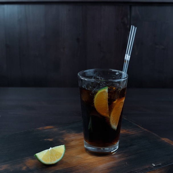 Το πρωτότυπο γευστικό κοκτέιλ με την προσθήκη πάγου, ουίσκι και κόκα-κόλα, ασβέστη φέτες στέκεται σε ένα ξύλινο τραπέζι σε ένα εστιατόριο. Η αρχική παρουσίαση των αλκοολούχων ποτών στη γραμμή. - Φωτογραφία, εικόνα