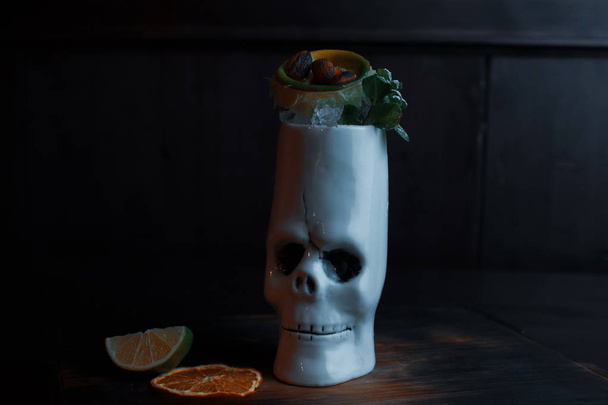 Beautiful tropical sweet cocktail with: tequila, white rum, vodka, martini, fruit pieces and mint leaves stands on a vintage table in a bar. Alcoholic fruit cocktail in the original white skull glass. - Photo, Image