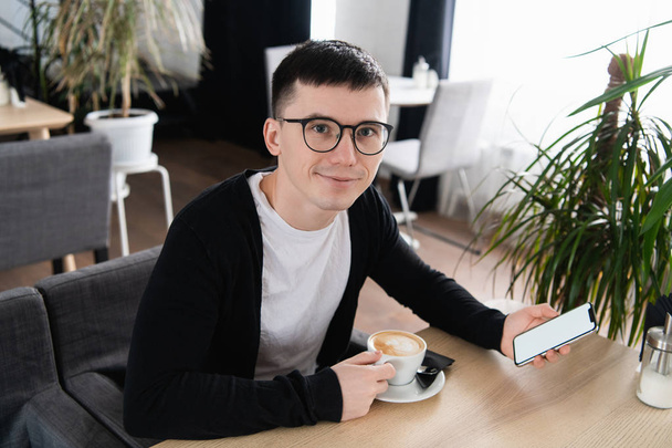 Happy smiling businessman or a student with a cup of coffee and smartphone in cafe. Place for your advertisment, app or logo. - Zdjęcie, obraz