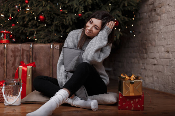 Nuori nainen tyylikkäässä vintage-puserossa mustissa housuissa valkoisissa sukissa istuu lattialla joulukuusen lähellä joulun kodikkaassa huoneessa lahjojen joukossa. Söpö tyttö ajattelee vapaapäiviä
. - Valokuva, kuva