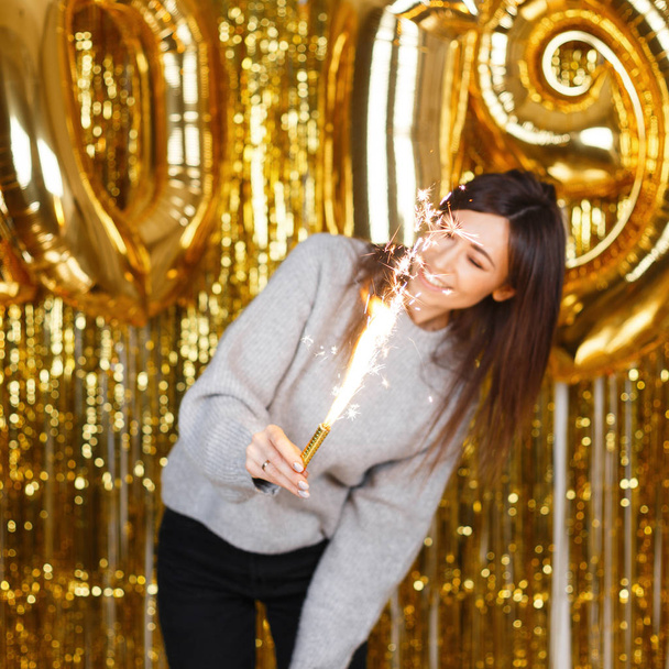 Young pretty woman in knitted sweater enjoy with fireworks in hands near the background of golden balloons 2019 and garlands in a festive studio. Girl is fun at the New Year's party. - Photo, Image
