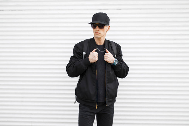 Ein junger, stylischer Hipster mit schwarzer Mütze und Sonnenbrille in stylischer Jeans, schwarzer Bomberjacke und einem Rucksack auf den Schultern steht auf der Straße in der Nähe einer weißen Metallwand. ernsthafter Modetyp. - Foto, Bild