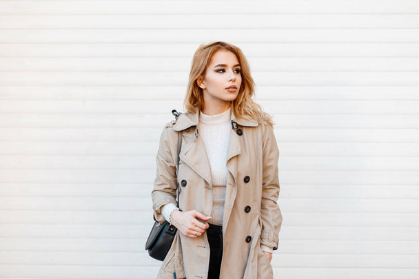 Szexi gyönyörű divatos fiatal nő, egy divatos tavaszi kabát, egy fehér pólót, és egy trendi fekete bőr táska fehér fém fal közelében áll. Európai stílusos lány. - Fotó, kép
