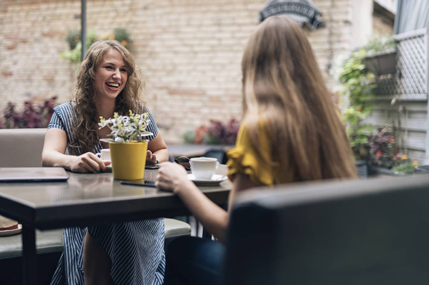 Stijlvolle jonge vrouwen met vriendelijke bijeenkomst met kopjes koffie terwijl zittend aan tafel en chatten - Foto, afbeelding