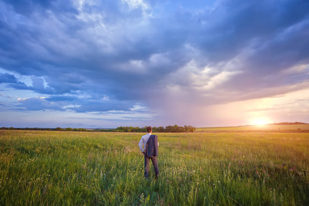 Biznesmen w elegancki garnitur z jego kurtkę wiszącą nad jego ramię stojący w polu patrząc na odległość pod majestatyczne wieczorne niebo z zachodzącego słońca. - Zdjęcie, obraz