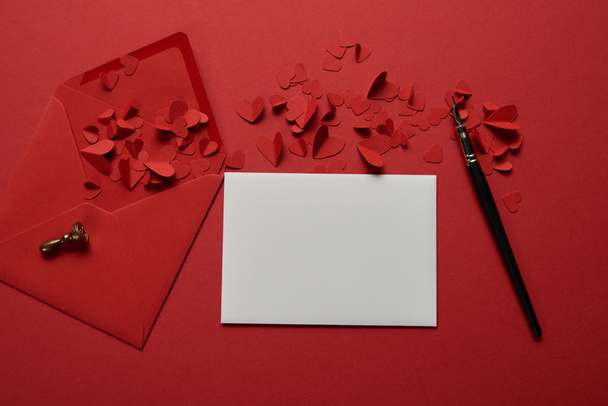 κάτοψη του άδειο λευκό γράμμα με φάκελο, κοπής χαρτιού καρδιές και στυλό πάνω σε κόκκινο φόντο - Φωτογραφία, εικόνα