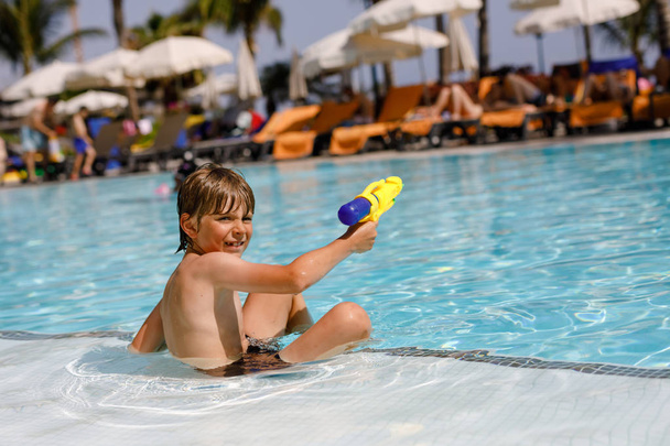 Happy little kid boy jumping in the pool and having fun on family vacations in a hotel resort. Healthy child playing in water with a water gun. Laughing running child splashing - Photo, Image