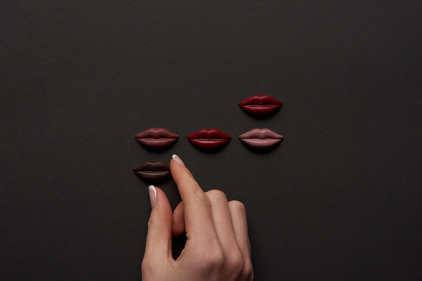 cropped view of female hand and multicolored chocolate lips on black background  - Photo, Image
