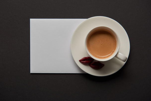 top view of cup with coffee, chocolate lips on saucer and empty white card on black background - Φωτογραφία, εικόνα