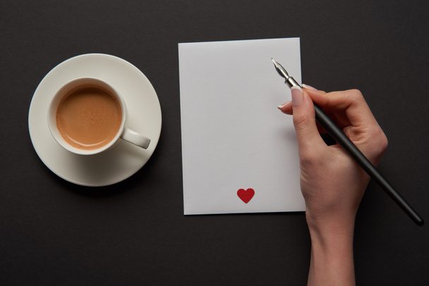 cropped view of woman writing on empty greeting card with red heart sign near cup of coffee - Photo, Image