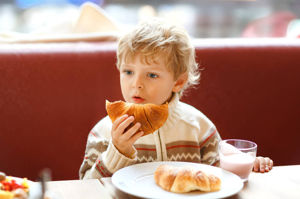 Cute healthy kid boy eating croissant and drinking strawberry milkshake in cafe. Happy child having breakfast with parents or at nursery. Vegetables, eggs as healthy food for children. - Photo, Image