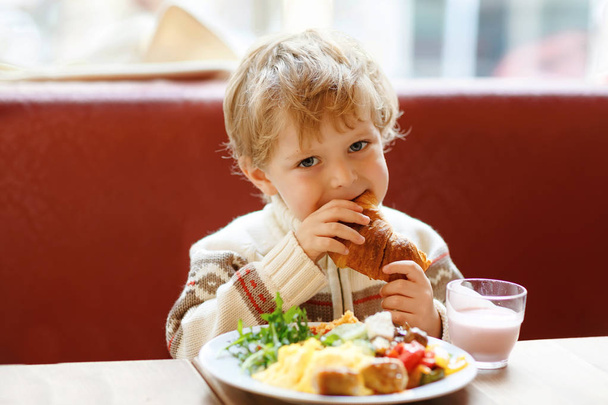 Chłopiec Cute dziecko zdrowe jedzenia rogalika i picia koktajl truskawkowy w kawiarni. Szczęśliwe dziecko posiadające śniadanie z rodzicami lub w przedszkolu. Warzywa, jaja, jak zdrowe jedzenie dla dzieci. - Zdjęcie, obraz